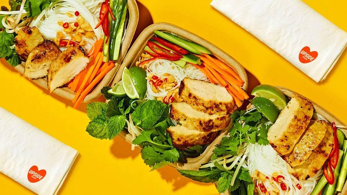 Three square paper containers of chicken chunks, greens and crudites with Upside Foods napkins nearby on a yellow background